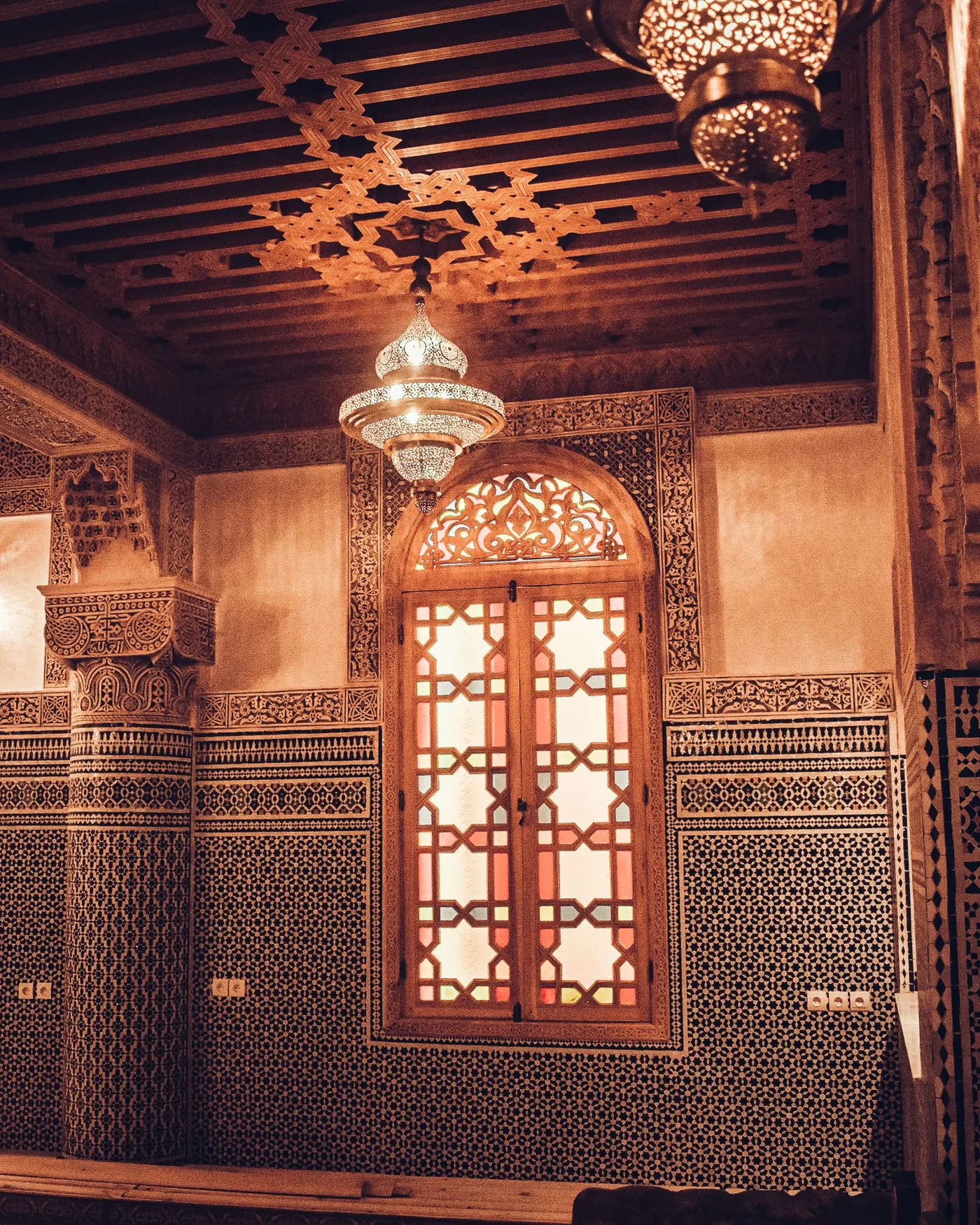 A Moroccan room with a carved cedar ceiling, a hanging lantern, a lit lattice window and tiled walls
