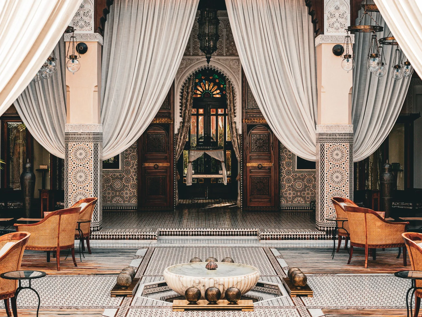 A Marrakesh courtyard with white drapes, tiled columns, a carved doorway and a mosaic floor around a marble fountain
