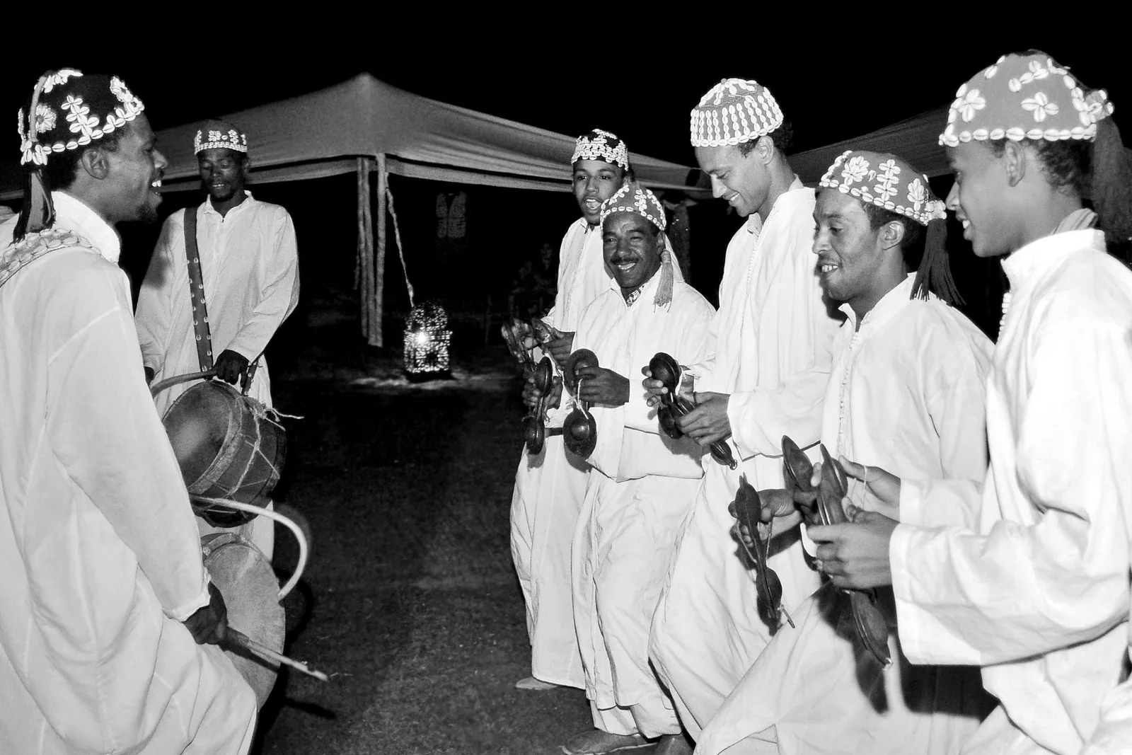 Black and white photograph of Gnawa musicians in white robes and cowrie-studded caps playing metal castanets at night