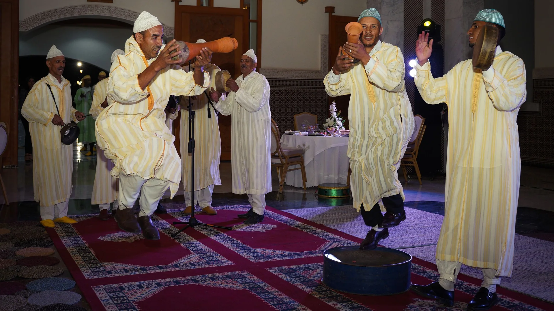Musicians in striped robes drumming and dancing on a red carpet in a Moroccan banquet hall, a laid table behind them