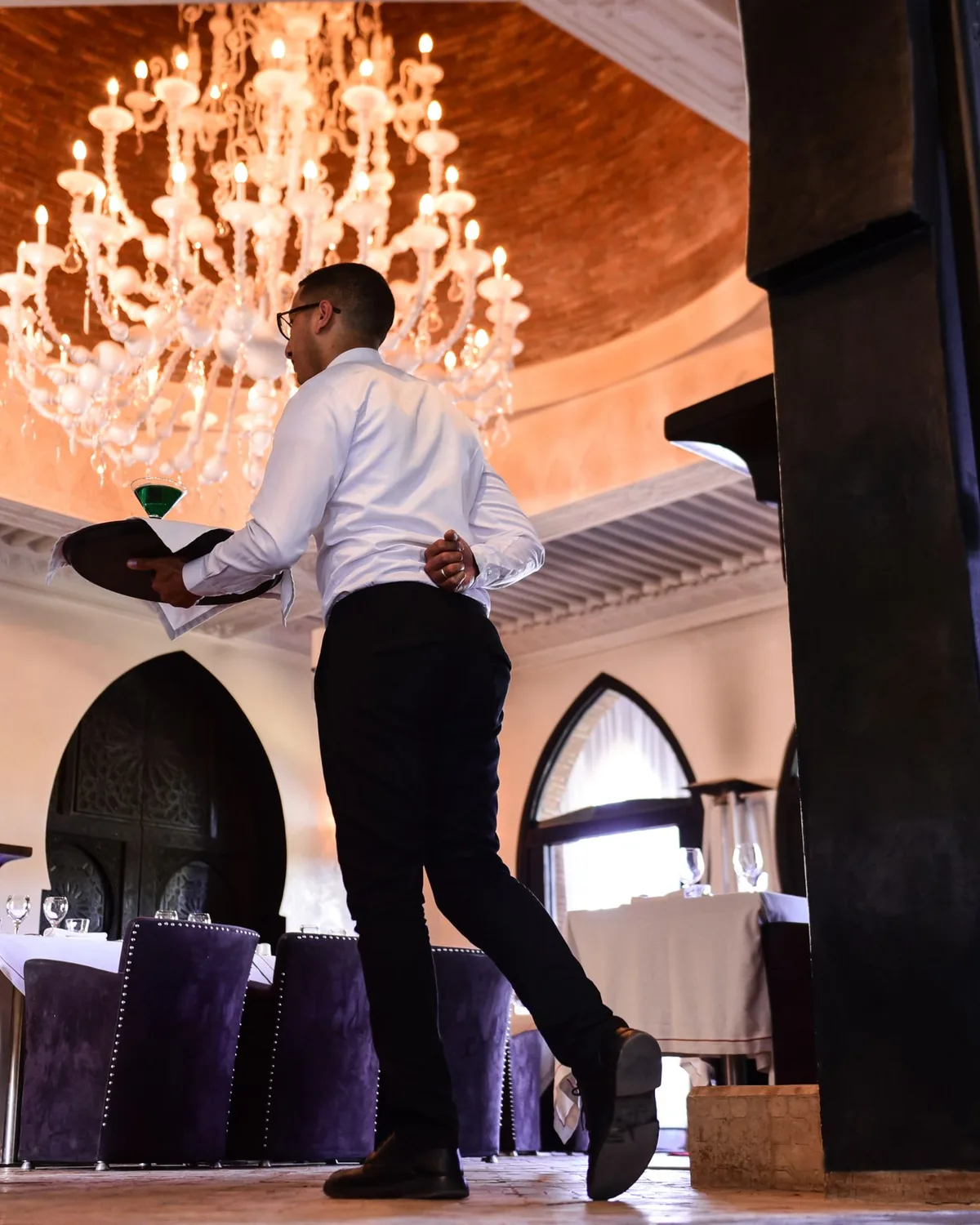A waiter carrying a tray beneath a large chandelier in a Marrakech dining room with pointed arches